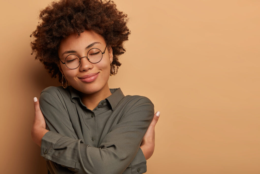 Tender woman dressed in dark shirt, embrasses herself, crosses arms over body, closes eyes with pleasure, has positive charming expression dreams about boyfriend hugges stands indoor has romantic mood